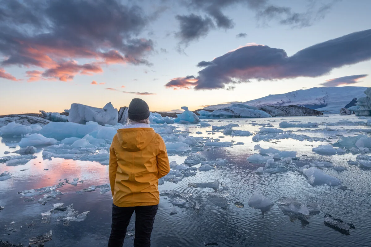 personne en ciré jaune face à une lagune avec des blocs de glace au coucher du soleil