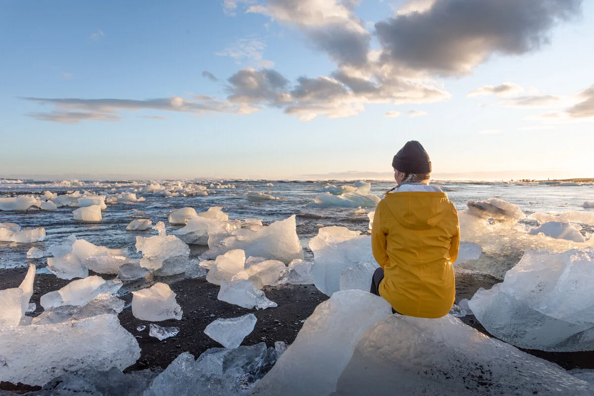 personne en veste jaune face assise sur un bloc de glace sur la Diamond Beach